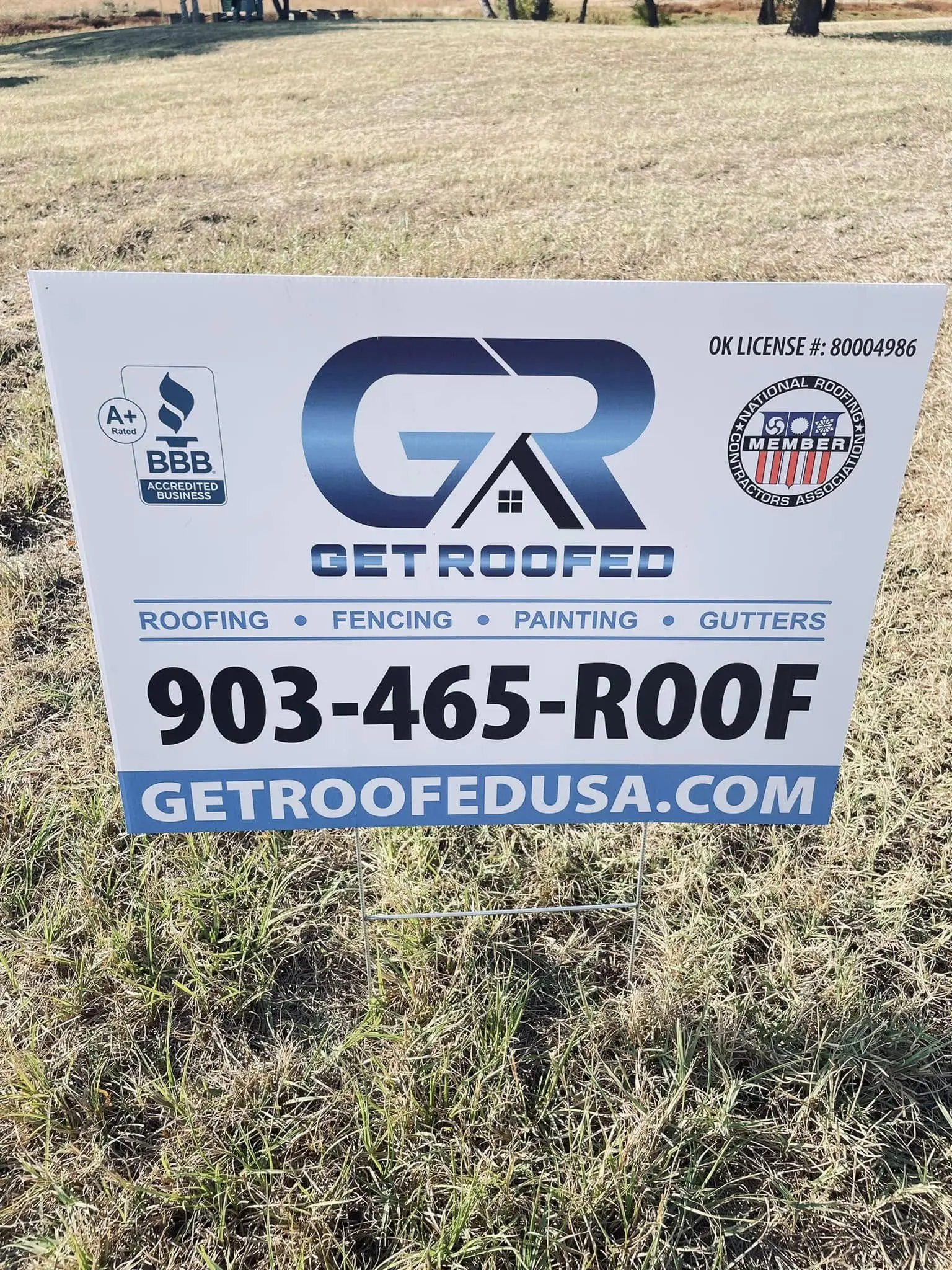 A sign in the grass advertising roofing and gutters.